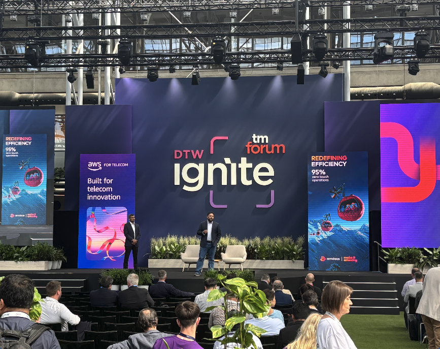 A speaker stands on a large conference stage in front of a dark blue backdrop with "DTW Ignite" and "TM Forum" logos.